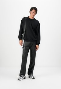 Jeune homme portant un sweat-shirt noir, un pantalon en cuir noir, des baskets argentées et noires, et un sac à bandoulière noir sur un fond blanc.