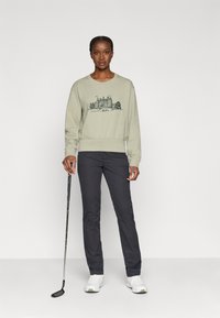 Gaiši zaļš džemperis ar melnu pils grafiku, saskaņots ar tumši pelēkiem biksēm un baltām tenisa kedām, turēt golfu nūju.