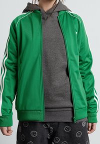 Persona con una chaqueta verde con cremallera sobre una sudadera gris y pantalones negros con patrones de caras sonrientes, de pie contra un fondo plano.