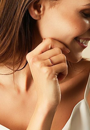 Dünner silberner Ring mit roten, rosa und klaren Edelsteinen. Am Zeigefinger getragen, die Hand unter dem Kinn ruhend vor einem neutralen Hintergrund.