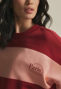 Punane ja roosa värvide blokeeringuga dressipluus ribitud kaelusega, millel on trükitud logo, mis ütleb "Paris Club d'Athlétisme". Pehme kangatunne.