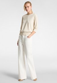 Maglione beige a maniche 3/4 con un leggero luccichio, abbinato a pantaloni bianchi a vita alta e a gamba larga. Il modello sta in piedi in modo casual con una mano in tasca.