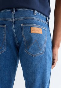 Povečava osebe, ki nosi modre Wrangler jeans, prikaz zadnjega žepa z usnjeno znamko in vezenim vzorcem.