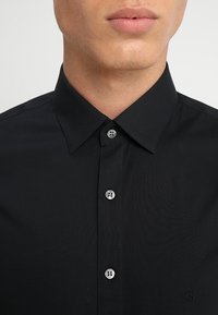 Zwarte button-up shirt van gladde stof, met een klassieke kraag, zes knopen en een klein logo aan de rechteronderzijde.