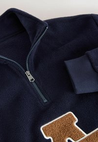 Pulover din fleece marin cu fermoar pe jumătate, manșete ribbate și o patch logo texturat, maro, înconjurat de alb. Material moale și confortabil, cu textură ridicată.