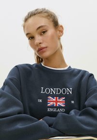 Mladá žena s vlasmi zviazanými dozadu, oblečená v námorníckej mikine s potlačou Union Jack a textom "Londýn UK Anglicko 98" na prednej strane, s prekríženými rukami.