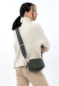 Szara skórzana torba na ramię o teksturowanej powierzchni, z regulowanym paskiem i detalami z zamkiem błyskawicznym, noszona przez osobę w kremowym swetrze i beżowych spodniach.