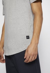 T-shirt cinza de algodão com mangas curtas, bainha arredondada e um pequeno rótulo de marca preto ao lado. Usado com calças escuras texturizadas.