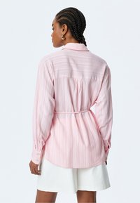 Camisa de rayas rosa claro con mangas largas, cintura ajustada y puño abotonado. Presenta un pliegue en la parte posterior y un dobladillo redondeado para un ajuste relajado.