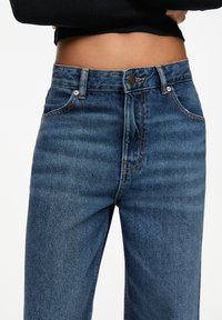 Vysoké modré džíny z denimu v klasickém pětikapsovém designu, s oranžovým prošíváním a kovovým zapínáním na knoflík.