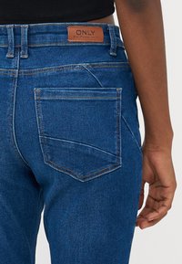 Modre denim hlače z visokim pasom, ravnim krojem in enim zadnjim žepom z vidnim šivom. Usnjen označevalec blagovne znamke na pasu.