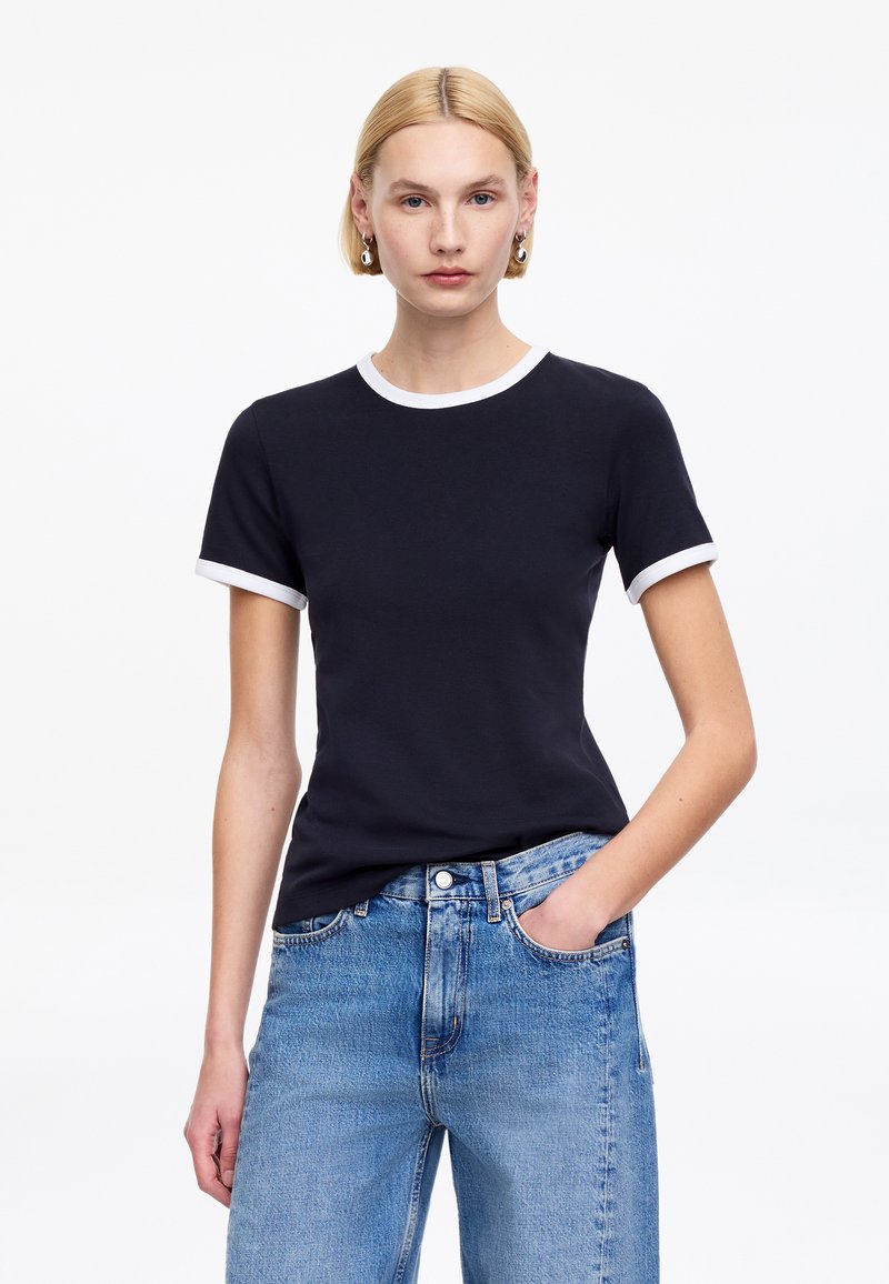 Jeune femme aux cheveux blonds courts portant un t-shirt noir à manches courtes avec un bord blanc et un jean bleu clair, debout avec une main dans la poche.