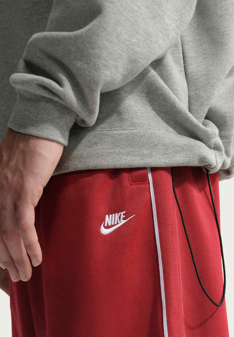 Main reposant sur le côté, portant un sweat gris et un pantalon Nike rouge avec un logo blanc et une bande blanche verticale.