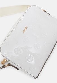 Bílá lesklá crossbody taška s vystouplými květinovými a písmennými vzory, se zlatým logem "JOOP!" na přední straně a béžovým popruhem.