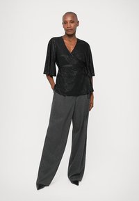 Zwarte, glanzende wikkeltop met korte mouwen, geknoopt in de taille, gecombineerd met grijze pantalon met wijde pijpen.