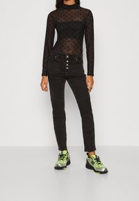 Top negro de malla de manga larga con un patrón sutil, cuello alto, combinado con pantalones ajustados negros con cierre de botones y acentos acanalados. Zapatillas verdes.