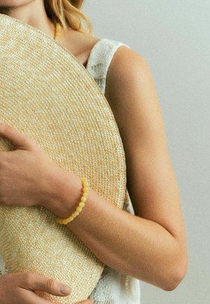 Frau trägt weißes ärmelloses Oberteil und hält großen, gewebten Strohhut, gelbes Perlarmband am Handgelenk vor einfarbigem Hintergrund sichtbar.