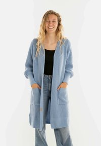 Glukkende vrouw met lang golvend blond haar draagt een lichtblauwe gebreide cardigan, een zwarte top en hoog getailleerde blauwe jeans, met haar handen in de zakken van de cardigan.