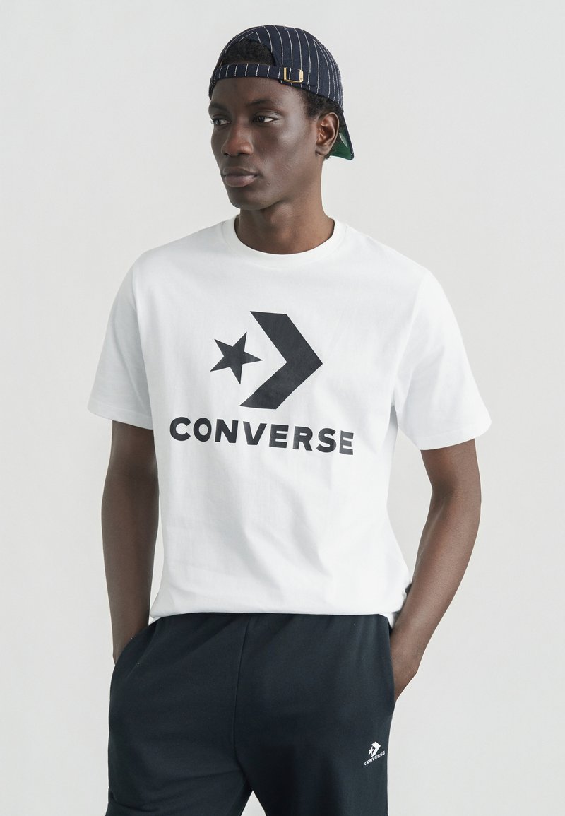 Hvid bomulds T-shirt med stort sort "CONVERSE" logo og grafisk stjerne, kombineret med sorte bukser og en marineblå kasket med tynde striber.