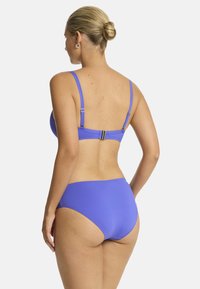 Una persona con un'acconciatura a donut indossa un bikini viola, mostrando il retro del top e il fondo a vita alta su uno sfondo semplice.