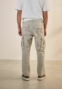 Pantalones cargo en un tono beige claro con bolsillos laterales, un diseño de pierna recta y un ajuste ligeramente relajado. Llevados con zapatos grises y una camiseta blanca.