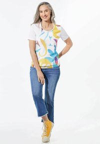 T-shirt met korte mouwen met een kleurrijk abstract patroon in geel, blauw en roze, gecombineerd met blauwe jeans en gele sneakers.