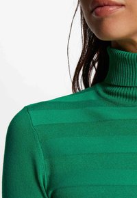 Suéter verde de cuello alto con textura acanalada y rayas horizontales. Presenta un diseño ajustado y una tela suave. Primer plano del área del cuello y el hombro.