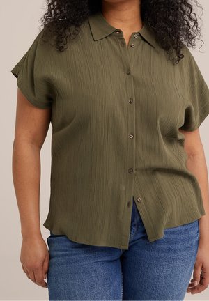 Kobieta w oliwkowo-zielonej, krótkim rękawie, koszuli na guziki z teksturowanej tkaniny i niebieskich dżinsach, stojąca na neutralnym tle.