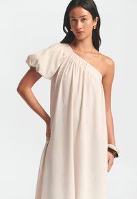 Vaalean beige-yksipuoleinen mekko, jossa on puffihihna, poimuinen kaula-aukko ja lentävä siluetti. Mallilla on leveä rannekoru yhdessä ranteessa.