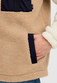 Chaqueta de forro polar beige con una superficie texturizada, que presenta un parche de tela oscura, un bolsillo lateral y un cuello de marfil en contraste. Diseño casual.
