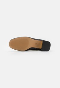 Suela de zapato de cuero negro con un tacón negro texturizado y goma, y una suela exterior marrón clara y lisa. Presenta un sutil logotipo en la suela.