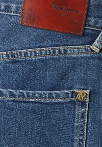  bolso de jeans de denim azul com costura amarela, um pequeno rebite de metal e uma etiqueta da marca Pepe Jeans London em couro marrom.