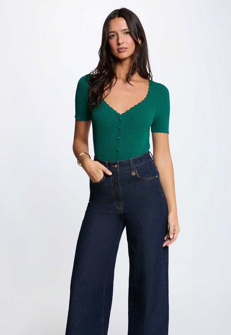 Woman with long dark hair wearing a green short-sleeve buttoned top and high-waisted dark blue wide-leg jeans, standing with one hand in pocket.
