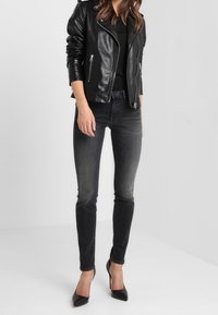 Kvinde iført sort læderjakke, sort top, slidte sorte skinny jeans og sorte spidse højhælede sko, stående mod en ensfarvet hvid baggrund.