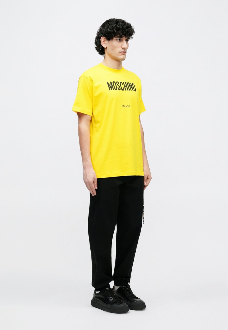 Gul bomuld T-shirt med "MOSCHINO"-logo i sort, kombineret med sorte bukser og sorte sneakers, båret af en mandlig model.