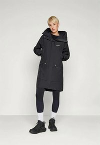 Giacca impermeabile nera con cappuccio, zip frontale e due tasche laterali. Caratterizzata da un taglio lungo e una texture liscia. Indossata sopra leggings neri e sneakers.