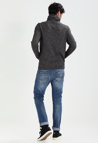Man som bär en mörkgrå polotröja, uppvikta blå jeans och svarta högskor, står med händerna på höfterna och vänder ryggen till.