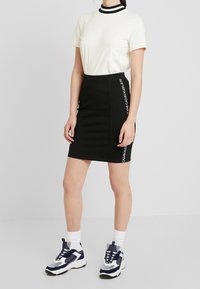 Černá pružná sukně s bočním logem, kombinovaná s bílým tričkem s černobílým pruhovaným límečkem, obutá s sportovními teniskami.