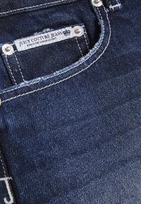 Σκούρο μπλε τζιν με εμφανές λευκό ράψιμο, μεταλλικά εξαρτήματα και ετικέτα με το εμπορικό σήμα «Juicy Couture Jeans».