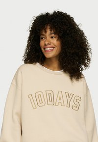 Sudadera beige con llamativos bordados en contraste de la palabra "10DAYS", con un corte relajado y textura suave.