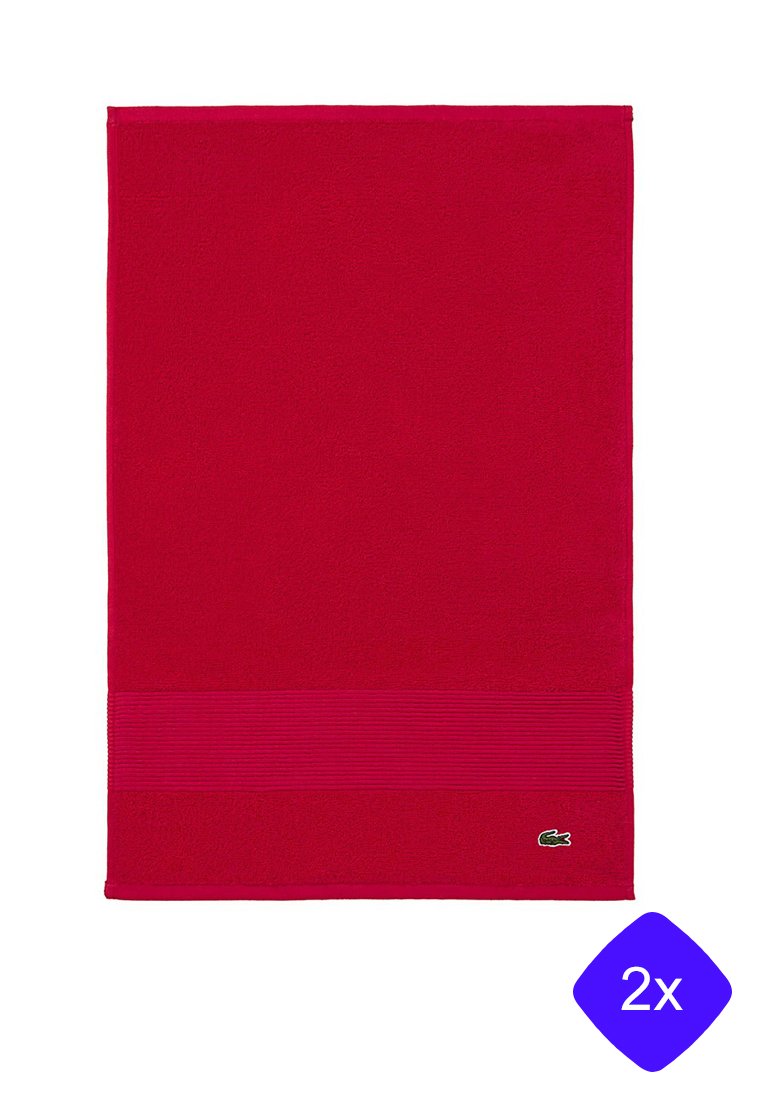 Toalla de baño de algodón roja con una franja texturizada horizontal en la parte inferior. Incluye un pequeño logo verde en la esquina inferior derecha. Forma rectangular.
