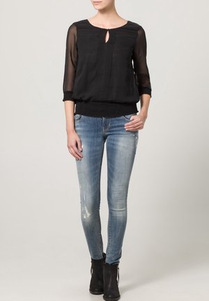 Zwarte doorzichtige blouse met een uitsnijding aan de voorkant, gecombineerd met lichtblauwe versleten skinny jeans en zwarte enkellaarsjes.
