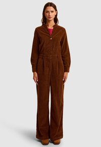 Brązowy kombinezon z końcówką z korduroy, z długim rękawem, z przodu zapinany na guziki, z elastyczną talią i szerokimi nogawkami. Gładka tekstura z prążkowanym wzorem.