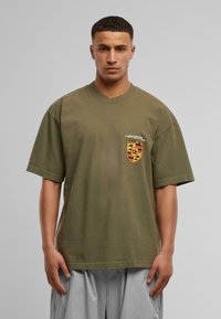 Olivengrøn oversized T-shirt i bomuldsstof med rund halsudskæring. Har et trykt emblem på venstre brystområde.
