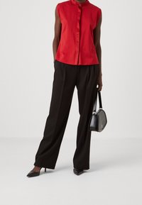 Camicetta rossa senza maniche con chiusura a bottoni, abbinata a pantaloni larghi neri. Il modello tiene una piccola borsa nera e indossa scarpe con tacco appuntito.