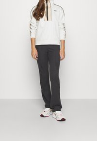 Sweat à capuche crème et beige avec un col montant, rayures noires, associé à un pantalon évasé côtelé foncé et des baskets blanches avec des touches de couleur.