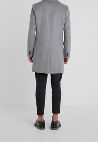 Abrigo de lana gris, corte entallado, con una abertura en la parte trasera, combinado con pantalones negros y zapatos negros brillantes. Diseño limpio y minimalista.