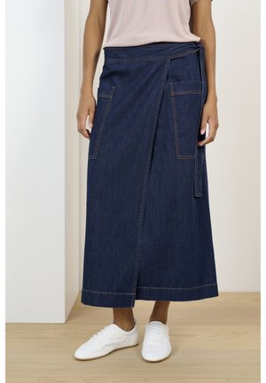 Kobieta w ciemnoniebieskiej spódnicy typu wrap z kieszeniami z przodu oraz białych sneakersach na sznurowadła, stojąca na jasnej drewnianej podłodze.