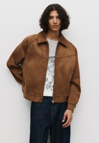 Veste en daim marron avec un large col, fermeture éclair à l'avant, poches latérales et coupe décontractée, associée à un sweat-shirt graphique gris et un jean foncé.