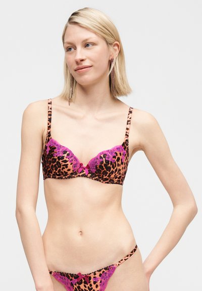 Szőke nő rózsaszín és fekete leopárdmintás melltartóban és hozzáillő, csipkedíszítéses alsóneműben, világosszürke háttér előtt állva.